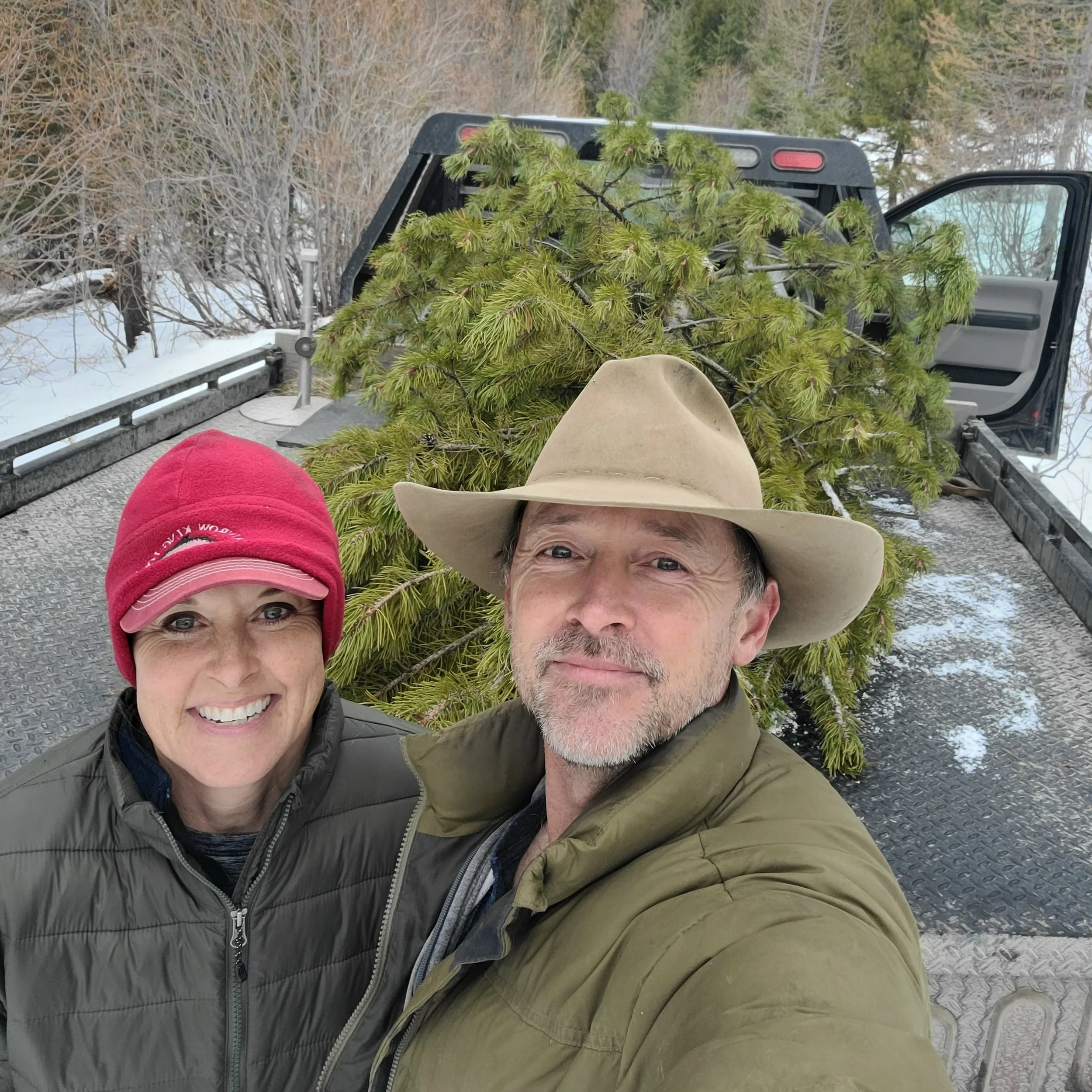 Brenda+Herron+-+20201219_142448 Couple posing for a selfie in front of a pickup truck loaded with a freshly cut pine tree, surrounded by snowy landscape and trees in the background.